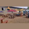 Alt Text Triangle sun shade sails suspended over an outdoor event seating area with wooden picnic benches and umbrella tables at Mistico Equestrian Centre.