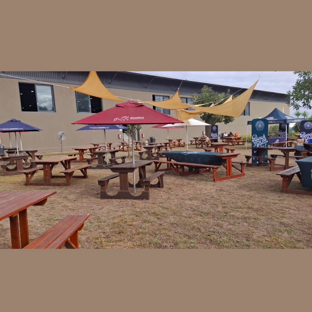 Alt Text Triangle sun shade sails suspended over an outdoor event seating area with wooden picnic benches and umbrella tables at Mistico Equestrian Centre.